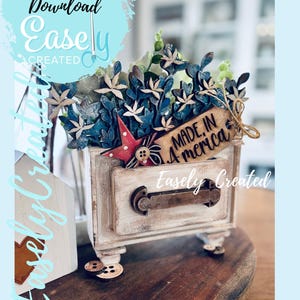 May include: A small, distressed white wooden chest with a drawer, adorned with blue and white floral accents, a red star, and a tag that reads "MADE IN AMERICA." The chest is on a wooden surface.