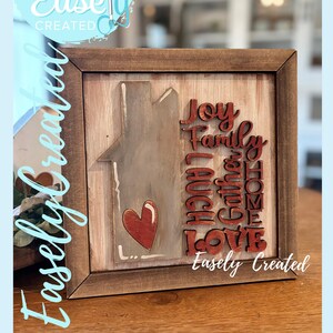Peut inclure: Panneau en bois encadré avec une silhouette de maison, un cœur rouge et les mots "Joy, Family, Gather, Home, Laugh, Love" dans un style rustique. Le panneau comprend également le texte "Easely Created" et "Digital Cut File".