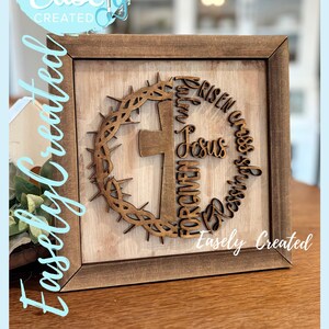 Peut inclure: Cadre en bois avec une croix, une couronne d'épines et les mots "Risen, Jesus, Forgiven, Blessings". L'œuvre d'art découpée au laser porte l'inscription "Easely Created". Le fond est en bois clair.