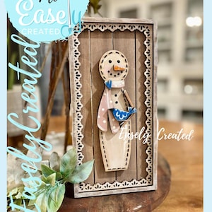 May include: Wooden snowman decoration with a light brown frame and a light blue background. The snowman has a carrot nose, black eyes, and a blue bird. The text "Digital Cut File Easily Created" is in the image.