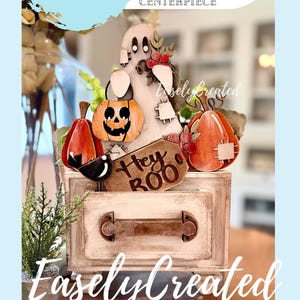 May include: A wooden Halloween-themed tiered tray decor set featuring a ghost, orange pumpkins, a black bird, and a brown tag with the text Hey BOO. The set includes a small rectangular wooden box with a handle and distressed white paint finish.