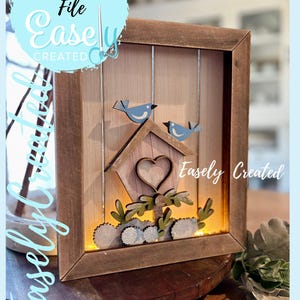 May include: A wooden framed wall art piece featuring a birdhouse with two blue birds perched on top. The birdhouse has a heart-shaped cutout and is surrounded by decorative foliage. The text "Easely Created" is visible.
