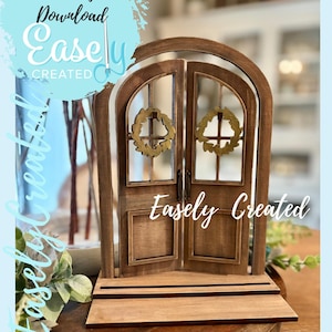 Op de afbeelding: Een kleine houten deur met een gewelfde bovenkant en twee deuren. Elke deur heeft een gouden krans. De tekst "Easely Created" staat op de deur. De achtergrond is lichtblauw met de tekst "Instant Digital Download Easely Created" in een cirkelvormig ontwerp.