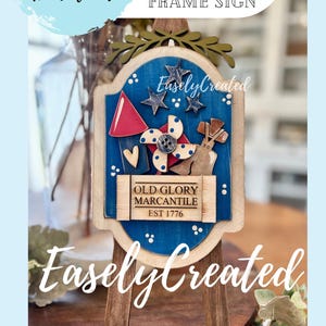 May include: A wooden sign with a blue background and patriotic decorations, including stars, a red firework, and a pinwheel. The sign reads "Old Glory Mercantile EST 1776" and "Easely Created". The sign is displayed on a wooden easel.