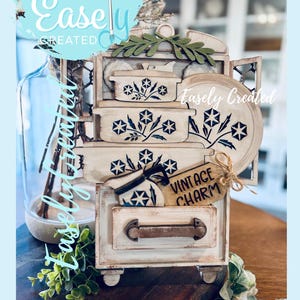 May include: A decorative wooden cabinet with a vintage charm theme. The cabinet is painted in cream and features drawers and shelves adorned with floral patterns in blue and white. The text "Vintage Charm" is on a tag.