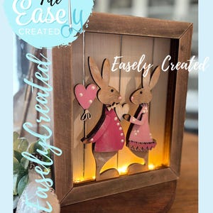 Puede incluir: Decoración de Pascua enmarcada en madera con dos figuras de conejos con abrigos rosas, sosteniendo un globo en forma de corazón y un corazón. El marco tiene una luz cálida en el interior, y el texto "Easely Created" es visible.
