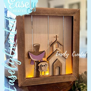 Puede incluir: Obra de arte enmarcada en madera con un diseño de cruz, iglesia y cordero. La cruz tiene un paño morado. La iglesia y el cordero son de madera clara. El texto "Easely Created" es visible en el marco y en un diseño circular.