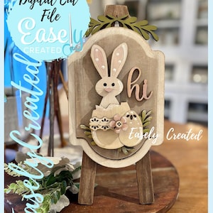 Puede incluir: Cartel de madera con temática de Pascua sobre un caballete pequeño. El cartel presenta un conejito saliendo de un huevo, con la palabra "hi" y el texto "Easely Created". El cartel está decorado con detalles florales y está sobre una base de madera.