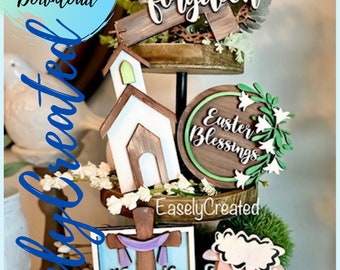 May include: A three-tiered wooden stand decorated with Easter-themed cutouts. The top tier features a wooden cross with the word "forgiven" on it, the middle tier has a white church cutout and a round wooden sign with the text "Easter Blessings", and the bottom tier has a square wooden sign with the text "He is Risen" and a white lamb cutout.