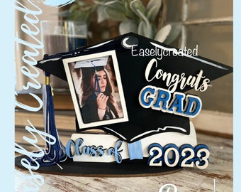 Könnte beinhalten: Eine schwarze Abschluss-Kappe mit einer blauen Quaste und einem weißen Rahmen für ein Foto. Die Kappe trägt die Worte "Congrats Grad" und "Class of 2023". Die Worte sind in Weiß mit blauen Umrissen. Die Kappe befindet sich auf einer Holzoberfläche.