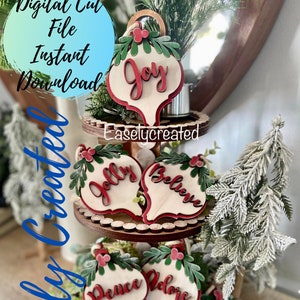 May include: A tiered wooden stand with six wooden ornaments shaped like Christmas ornaments. Each ornament has a different word on it: "Joy", "Jolly", "Believe", "Peace", "Adore", and "Merry". The ornaments are painted white with red lettering and have green leaves and red berries on top.