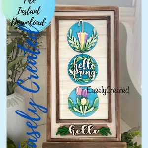 May include: A wooden sign with three round decorations. The top decoration is a blue circle with a green wreath and a yellow and purple umbrella. The middle decoration is a blue circle with a green wreath and the text "hello spring". The bottom decoration is a blue circle with a green wreath and three pink, purple, and yellow tulips. The sign is on a wooden easel.
