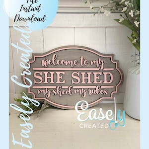 May include: A wooden sign with a pink border and the text "Welcome to my SHE SHED my shed my rules". The sign is on a white wooden surface with a plant in the background.