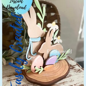 Pode incluir: Um kit de artesanato de coelho da Páscoa de madeira com dois coelhos, flores, folhas, ovos e uma cenoura. O kit está sobre uma base de fatia de madeira. O texto "Easilycreated" é visível na base.