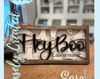 May include: A wooden sign with a white background and a black inscription that reads "Hey Boo" with a ghost design. The sign is framed in a dark brown wood frame. The sign is on a wooden surface. The text "Easlycreated" is in the bottom right corner of the sign.