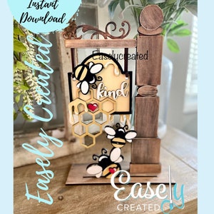 May include: A wooden sign with a bee and honeycomb design. The sign says "Be Kind". The sign is attached to a wooden post with a decorative top.