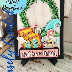 Op de afbeelding: Een houten gnome met een blauwe en witte golfhoed, die een bord vasthoudt met de tekst "beach bum". De gnome is omringd door schelpen, slippers en een groene krans van bladeren. Het bord staat op een roze en zwarte standaard met de tekst "home sweet home" erop.