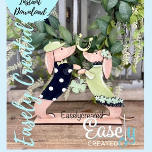 May include: Two wooden dachshund figurines, one wearing a black and white polka dot dress and a green hat, the other wearing a green dress with green shamrocks, holding a gold horseshoe. The figurines are standing on a wooden base with the text "Easlycreated" printed on it.