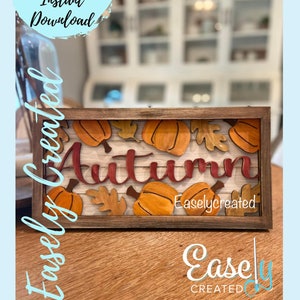 May include: A wooden framed sign with the word "Autumn" in red lettering. The sign is decorated with orange pumpkins and brown leaves. The sign is on a wooden surface. The text "Easlycreated" is visible in the bottom right corner of the sign.