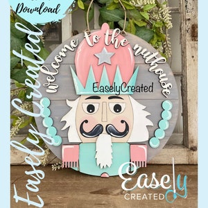 May include: A wooden round sign with a nutcracker design. The sign is painted in shades of pink, blue, and gray. The text "Welcome to the Nut House" is written in white cursive lettering. The sign is decorated with a star and beads.