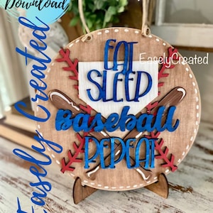 May include: A wooden round sign with the text "Eat Sleep Baseball Repeat" in blue lettering. The sign is decorated with a baseball mitt, two baseball bats, and red stitching. The sign is on a small wooden stand.