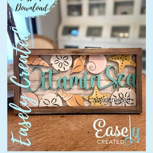 May include: A wooden framed sign with the text "Vitamin Sea" in teal blue lettering. The sign is decorated with seashells, starfish, and sand dollars in shades of orange, yellow, and pink. The sign is on a wooden surface.
