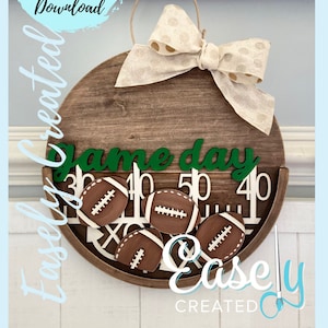 May include: A wooden round sign with a brown background and a green "Game Day" inscription. The sign has a white and brown football design with a white and brown bow on top. The sign is hanging on a white wall.