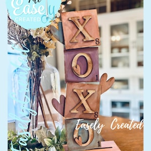 以下が含まれることがあります： 素朴な仕上がりの木製XOXOブロックセット。ブロックは茶色、紫、青緑色で塗装されており、文字と数字が切り抜かれています。「Easely Created」の文字も表示されています。
