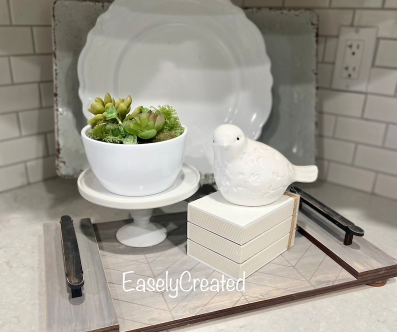 May include: A white ceramic bird figurine sits on a stack of white boxes on a wooden tray with a gray and white geometric pattern. A white ceramic planter with a succulent plant sits on a white pedestal on the tray. The tray has a black metal handle. The text "EaslyCreated" is printed on the tray.