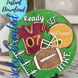 May include: A wooden football-themed sign with a green background, featuring a maroon jersey with the number 07, a yellow helmet, and a brown football. The words "Ready, Set, Hike" are displayed.