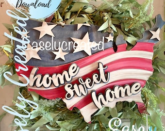 May include: A wooden cutout of the United States in the shape of a map with red, white, and blue stripes and stars. The words "home sweet home" are written in a cursive font on the cutout. The cutout is surrounded by green foliage.
