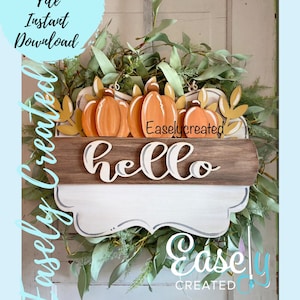 Può includere: Un cartello di legno con la parola "hello" in caratteri bianchi su sfondo marrone. Il cartello è decorato con una ghirlanda di foglie verdi e zucche arancioni. Il testo "Easlycreated" è visibile sul cartello.