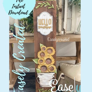 Peut inclure: Une pancarte en bois avec une finition brune rustique présentant une pancarte blanche avec le texte "Hello Sunshine" et un seau rempli de tournesols. La pancarte est décorée de feuilles vertes et le texte "Easlycreated" est imprimé sur la pancarte.