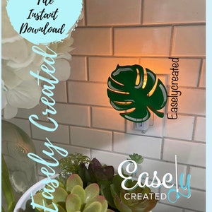 May include: A green monstera leaf shaped night light plugged into an electrical outlet. The light is on and illuminating the leaf. The text "Easley Created" is visible in the image.
