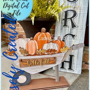 May include: A white wooden wheelbarrow filled with orange and white pumpkins and fall leaves. The wheelbarrow has a wooden base and a sign that says "hello fall".