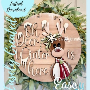 May include: A wooden round sign with the text "Oh Deer! Winter is here" and a painted reindeer with a red scarf and a snowflake on its head. The sign is surrounded by green leaves.