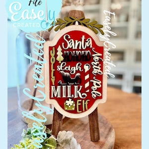 May include: A wooden Christmas sign with a red background and white lettering that reads "Santa", "Ho Ho Ho", "sleigh", "North Pole", "Milk", and "Elf". The sign is on a small wooden easel. The words "Digital Cut File" and "Easely Created" are also visible.