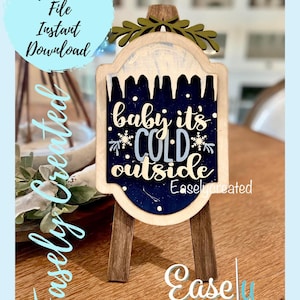May include: A wooden sign with a blue background and white lettering that reads "baby it's cold outside". The sign is decorated with snowflakes and a green leafy garland. The sign is on a wooden easel.