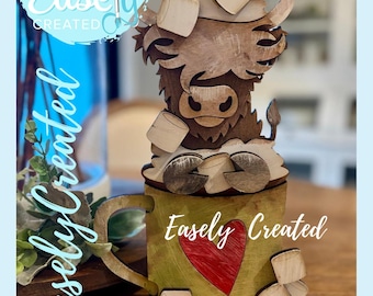 May include: Wooden Highland cow figurine with marshmallows, sitting atop a green mug with a red heart. The mug and cow are made of wood. The text "Easely Created" is on the mug and in the background. The words "Digital Cut File" are in the background.