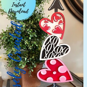 May include: A wooden Valentine's Day decoration with three layered hearts. The top heart is white with red hearts, the middle heart is black with white "XOXO" lettering, and the bottom heart is red with pink polka dots. The hearts are attached to a black arrow and a black base.