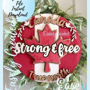 Puede incluir: Un letrero de madera rojo, blanco y marrón con las palabras "Canadá", "Strong & Free" y "True North" en él. El letrero está decorado con una hoja de arce y una corona de hojas verdes.