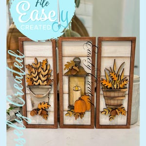 May include: Three wooden framed wall art pieces with fall-themed designs. Each piece features a light-colored background with a wooden frame. The designs include fall foliage, pumpkins, and a lantern. The text "Digital Cut File Easely Created" is visible.