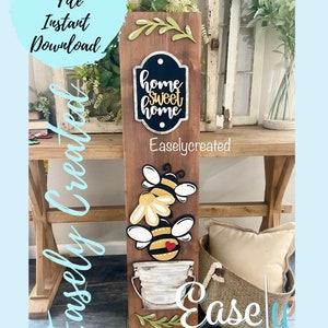 May include: A wooden sign with a bee theme. The sign features a bucket, two bees, a flower, and a chalkboard sign that says "home sweet home".