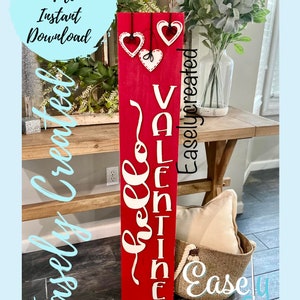 May include: Red wooden sign with white lettering that reads "Hello Valentine". The sign is decorated with three white hearts with stitching detail. The sign is leaning against a wooden table.