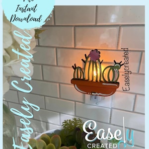 Puede incluir: Una luz nocturna de cactus de madera con una flor rosa en la parte superior, enchufada a una toma de corriente. La luz está encendida y brillando. El texto "Easley Created" es visible en la esquina inferior derecha de la imagen.