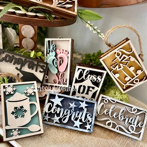 May include: A collection of wooden signs with various sayings, including "Congrats", "He or She", "Class of", "Celebrate", and "Congrats". The signs are decorated with different colours and designs, such as stars, snowflakes, and footprints.