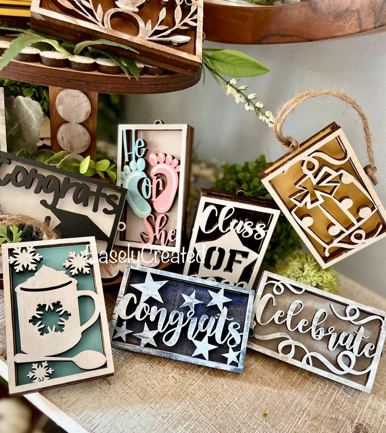 May include: A collection of wooden signs with various sayings, including "Congrats", "He or She", "Class of", "Celebrate", and "Congrats". The signs are decorated with different colours and designs, such as stars, snowflakes, and footprints.