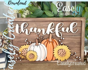 Könnte beinhalten: Ein Holzschild mit dem Wort "thankful" in weißen Buchstaben. Das Schild ist mit drei Kürbissen und Sonnenblumen verziert, alle in Orange-, Weiß- und Gelbtönen.