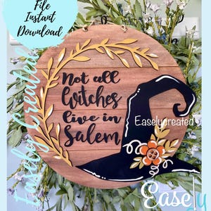 Puede incluir: Un letrero redondo de madera con el texto "Not all witches live in Salem" y un sombrero de bruja negro con detalles florales naranjas y amarillos. El letrero está rodeado de una corona de hojas verdes.