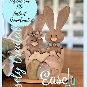 May include: Two wooden bunny rabbits in an egg-shaped nest with a heart on the base. The bunnies are painted brown with white accents and have brown eyes. The eggs are painted in shades of green and pink. The base is painted brown. The text "Easly Created" is on the base.